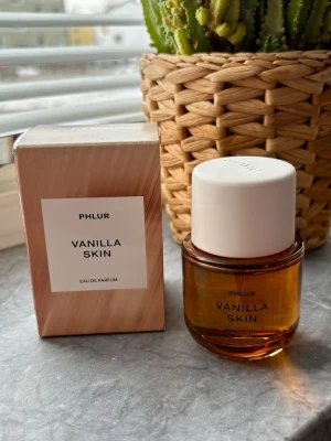 Phlur Vanilla Skin Eau de Parfum - Phlur Vanilla Skin Eau de Parfum kommer i en rund, bärnstensfärgad glasflaska med vit kork och stilren etikett. Doften har en varm och söt vaniljkaraktär. Förpackningen är elegant med mjuka beige toner och minimalistisk design. 50ml, använd enstaka gång