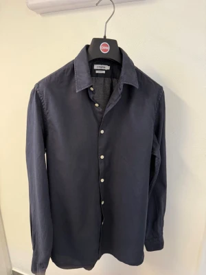 Mörkblå skjorta från J.Lindberg - Stilren mörkblå skjorta från J.Lindberg med klassisk krage och vita knappar. Skjortan har lång ärm och är tillverkad i bomull för en bekväm känsla. Perfekt för dig som gillar enkel och tidlös stil.