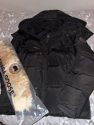 Svart Canada Goose Wyndham - Säljer en svart dunjacka från Canada Goose med avtagbar pälskant till huvan. Jackan har klassisk quiltad design, två sidofickor och tydlig logotyp på ärmen. Perfekt för kalla vinterdagar och riktigt varm tack vare dunfyllningen.