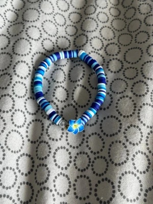 Blått pärlarmband med blomma - Snyggt armband med platta pärlor i olika blå och vita nyanser. Armbandet har en detalj i form av en blå och gul blomma i mitten samt en silverfärgad pärla bredvid. Perfekt accessoar för att addera färg till din outfit.
