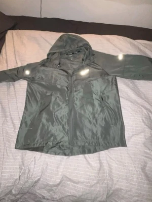 Grå Nike Miller  - Snygg grön vindjacka med huva och dragkedja framtill. Jackan har två sidofickor med dragkedja och justerbar dragsko i huvan. Materialet är tunt och lätt, perfekt för blåsiga dagar. Reflekterande detaljer på axlarna för extra synlighet.