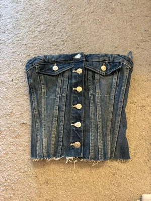 Denim korsett med knappar från Zara - Cool denim korsett från Zara med knappar framtill och rå fransad nederkant. Toppen har fickdetaljer och smock baktill för extra komfort. Perfekt för dig som vill ha en edgy och trendig look.