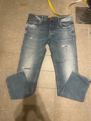 Blå slitna jeans med raka ben jack and Jones  - Säljer ett par blå jeans med raka ben och slitna detaljer på framsidan. Jeansen har klassisk femficksmodell och ljus tvätt med snygga slitningar för en avslappnad look. Perfekta för dig som gillar en casual och trendig stil.