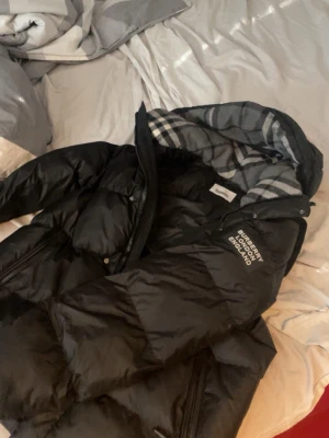 Svart pufferjacka från Burberry - Säljer en svart pufferjacka från Burberry med klassiskt rutigt foder i grått och vitt. Jackan har dragkedja, hög krage och logga tryckt på bröstet. Perfekt för kalla vinterdagar och går att ta av armarna och göra jackan till en väst. riktigt snygg till streetwear-looken.
