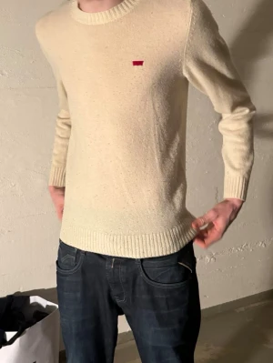 Beige stickad tröja från Levi's - Säljer en stilren beige stickad tröja från Levi's med rund halsringning och diskret röd logotyp på bröstet. Tröjan har långa ärmar och ribbade muddar vid ärmslut och nederkant. Perfekt för dig som gillar enkel och klassisk stil. Modellen på bilden är 187 cm och väger 70 kilo. Storleken är xs men passar även för S och vissa fall M