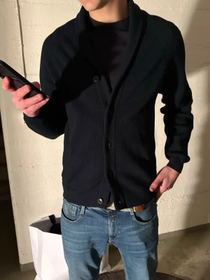 Mörkblå ribbad kofta med sjalkrage - Snygg mörkblå kofta med ribbad struktur och sjalkrage. Koftan har knäppning framtill med svarta knappar och långa ärmar. Perfekt att slänga över en t-shirt för en chill men stilren look. Modellen är 188 cm lång och väger ca 76 kilo