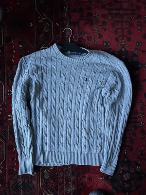 Ljusgrå kabelstickad tröja Polo Ralph Lauren - Säljer en ljusgrå kabelstickad tröja från Polo Ralph Lauren med rund halsringning och diskret broderad logga på bröstet. Tröjan har långa ärmar och ribbade muddar vid ärmslut och nederkant. Perfekt för dig som gillar klassisk och stilren look.