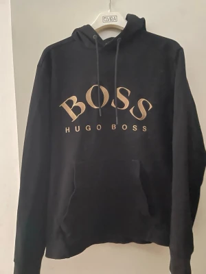 Svart hoodie från Hugo Boss - Snygg svart hoodie från Hugo Boss med stor guldfärgad BOSS-logga på bröstet. Klassisk huva med snörning och stor magficka framtill. Perfekt för dig som vill ha en stilren och trendig look.
