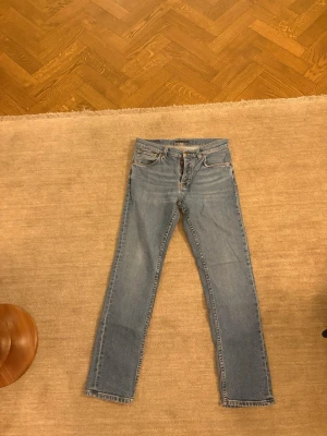 Ljusblå raka jeans från Nudie Jeans - Säljer ett par klassiska ljusblå jeans från Nudie Jeans med raka ben och femficksmodell. Jeansen har snygga kontrastsömmar och ikoniska broderade bakfickor. Tillverkade i slitstark denim med en tidlös look. Grim Tim 30/30