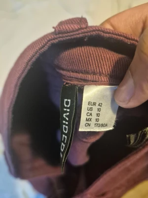 Vinröda utsvängda byxor från Divided - Säljer ett par lila utsvängda byxor från Divided. Byxorna har två bakfickor med lock och knappdetaljer. Materialet är mjukt och stretchigt, perfekt för en trendig och bekväm look.