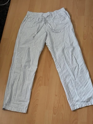 Randiga linnebyxor från H&M - Säljer ett par vita linnebyxor från H&M med tunna blå ränder. Byxorna har rak passform, resår i midjan och dragsko för skön och avslappnad stil. Perfekta för sommaren och tillverkade i ett lätt och luftigt material.
