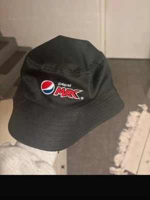 Svart bucket hat Pepsi Max - Säljer en svart bucket hat med broderad Pepsi Max-logga på framsidan och texten 'CHALLENGE YOUR TASTE' på baksidan. Hatten är gjord i ett slitstarkt tyg och har en klassisk form med bred skärm. Perfekt för dig som gillar streetwear och vill sticka ut.