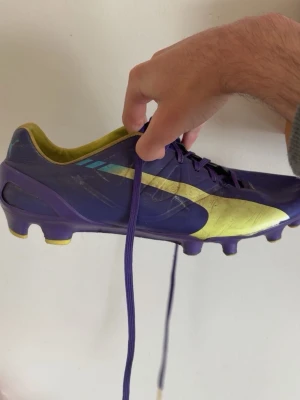 Puma fotbollsskor - Säljer ett par snygga fotbollsskor från Puma i lila och gult. Skorna har klassisk låg profil, snörning och mönstrad sula för bra grepp på planen. Tillverkade i syntetmaterial med coola detaljer och stor Puma-logga på sidan. Perfekta för dig som vill sticka ut på fotbollsplanen.