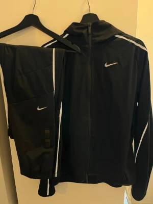 Nike Nocta svart träningsset med huva - Snyggt svart träningsset från Nike med jacka och byxor. Jackan har huva, hel dragkedja och vita detaljer längs ärmar och axlar. Byxorna har vita ränder och små Nike-loggor. Perfekt för löpning eller gym. Materialet är lätt och funktionsanpassat. Nocta , som ny 