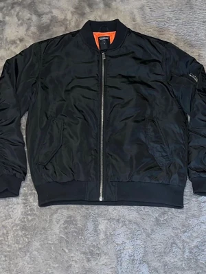 Klassisk bomberjacka ”ART 3)” - Trendig bomberjacka med det ikoniska orangea fodret. Jackan har ficka på ärmen och ribbade muddar. • Färger: Olivgrön (Oanvänd)