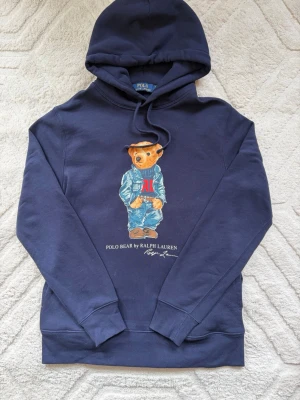 Marinblå hoodie Polo Bear Ralph Lauren - Snygg marinblå hoodie från Ralph Lauren med den ikoniska Polo Bear-trycket framtill. Tröjan har huva med dragsko och ribbade muddar vid ärmslut och nederkant. Perfekt för en avslappnad och trendig stil.