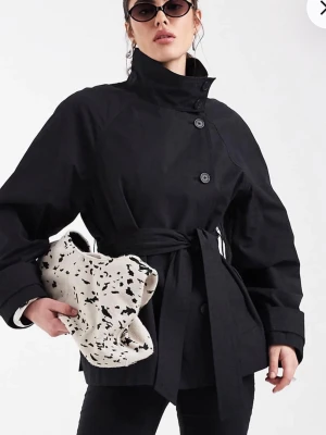 Svart trenchcoat med knytskärp - Stilren svart trenchcoat med hög krage, stora knappar och knytskärp i midjan. Jackan har vida ärmar och en modern, boxig passform. Perfekt för dig som gillar minimalistisk design och vill ha en cool look. Materialet ser ut att vara bomull eller polyester. Helt ny med prislapp. 