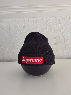 Svart ribbstickad mössa - Svart ribbstickad mössa med klassisk röd box-logo framtill. Mössan har uppvikt kant och är tillverkad i mjukt material som håller dig varm. Perfekt för dig som vill ha en streetwear-look och gillar ikoniska märken.