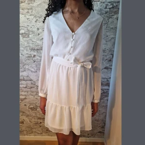 Vit klänning med knytband - Söt vit kortklänning med v-ringning, knappar framtill och ett knytband i midjan. Klänningen har långa, transparenta ärmar och volang nedtill för en romantisk look. Perfekt för sommarens alla tillfällen. Något off white. Stl 32/34.