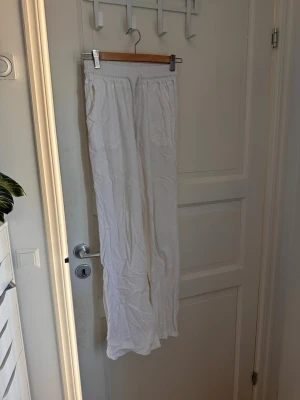 Vita vida linnebyxor med resårmidja - Säljer ett par vita linnebyxor med avslappnad passform och resår i midjan. Byxorna har raka ben och är tillverkade i ett lätt och luftigt linnematerial, perfekta för varma dagar. Inga synliga detaljer eller loggor.