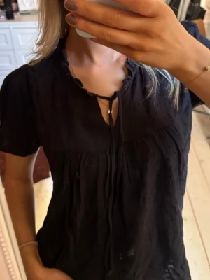 Svart blus med knyt och kort ärm - Svart blus med korta ärmar och diskret struktur i tyget. Blusen har en liten volangkant vid halsen och knytband framtill som ger en söt detalj. Passar perfekt till både jeans och kjol för en trendig look.