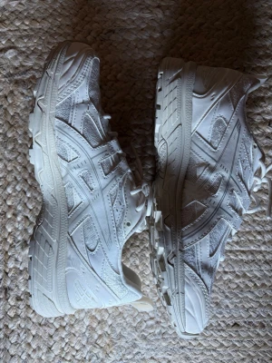 Vita Asics sneakers med meshdetaljer - Säljer ett par vita Asics sneakers med snygga meshdetaljer och mönstrade paneler. Skorna har en chunky sula och klassisk snörning. Perfekta för dig som gillar sportig stil och vill ha bekväma skor till vardags. Materialet är en mix av syntet och mesh.