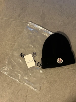 MONCLER MÖSSA - Snygg svart mössa från Moncler med uppvikt kant och klassisk Moncler-logga framtill. Tillverkad i mjukt stickat material som håller dig varm. Perfekt accessoar för kalla dagar och enkel att matcha med olika outfits.