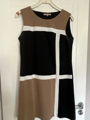 Beige och svart klänning från Anna Field - Snygg ärmlös A-linjeformad klänning från Anna Field i beige och svart med vita geometriska linjer. Klänningen har en rak och enkel form som ger ett stilrent intryck. Perfekt för dig som gillar grafiska mönster och klassiska färger.
