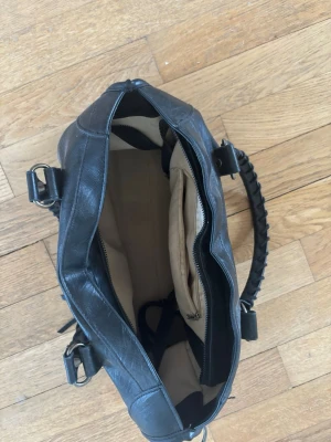 Svart handväska med flätade handtag - Snygg svart handväska i konstläder med flätade handtag och silverfärgade nitar. Väskan har ett stort huvudfack med ljus insida och innerficka med dragkedja. Perfekt för dig som vill ha plats för allt du behöver under dagen.