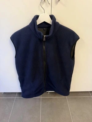 Mörkblå fleeceväst med dragkedja stl L - Mysig mörkblå fleeceväst med hel dragkedja framtill och hög krage. Västen har svarta kantband vid ärmhålen och två sidofickor. Perfekt att slänga på sig över en hoodie eller tröja för extra värme. Enkel och stilren design utan synligt märke.
