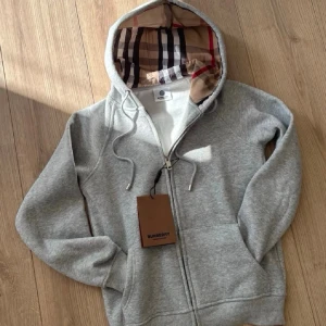 🩶Grå hoodie med design🩶 - Säljer en grå hoodie från Burberry med dragkedja och klassisk rutmönstrad huva i beige, svart och rött. Hoodien har snören i huvan och två fickor framtill. Perfekt för dig som vill ha en stilren och exklusiv look. Storlek xs passar även personer som är från 150-165 cm. Tar emot bud. Aldrig använd 