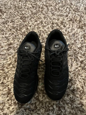 Nike Air Max Plus svarta sneakers - Säljer ett par Nike Air Max Plus sneakers i helsvart färg med mesh och syntetmaterial. Skorna har en chunky sula med synliga Air-bubblor och klassisk swoosh på sidan. Perfekt för dig som gillar en sportig och modern look. Helt onvända.