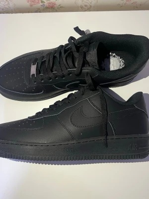 Nike Air Force 1 Low helsvarta sneakers - Säljer ett par klassiska Nike Air Force 1 Low sneakers i helsvart läder. Skorna har perforerad tå, kraftig sula med Air-dämpning och ikonisk Swoosh på sidorna. Perfekta för dig som gillar en clean och tidlös look.