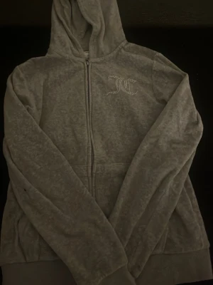 Grå velour hoodie från Juicy Couture - Säljer en grå velour hoodie från Juicy Couture med dragkedja framtill och huva. På bröstet finns Juicy Couture-loggan i glittrande stenar som ger en lyxig känsla. Hoodien har långa ärmar och mjukt material som känns skönt mot huden.
