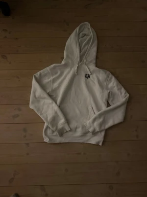 Vit hoodie med svart logotyp - Säljer en vit hoodie med svart broderad EBB-logotyp på bröstet. Hoodien har en stor huva med dragsko och en klassisk känguruficka framtill. Materialet är mjukt och bekvämt, perfekt för chill dagar. Passar dig som gillar stilren design och streetwear.