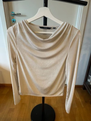 Champagnefärgad topp från Vero Moda - Säljer en snygg champagnefärgad topp från Vero Moda med draperad halsringning och långärmad design. Materialet har en lätt glansig finish som ger ett lyxigt intryck. Perfekt att matcha med jeans eller kjol för en cool look.