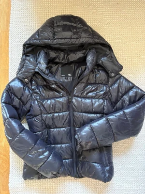 Svart pufferjacka från 157 med huva - Snygg svart pufferjacka från 157 med quiltad design och stor huva. Jackan har dragkedja framtill, långa ärmar och är tillverkad i glansig polyester. Perfekt för kalla dagar när du vill hålla dig varm och se cool ut.