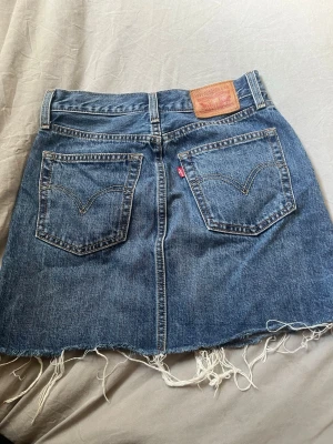 Levi's blå jeanskjol med fransig kant - Cool blå jeanskjol från Levi's med klassiska bakfickor och rå, fransig nederkant. Kjolen har knappstängning framtill och tydliga sömmar. Perfekt för dig som gillar en avslappnad och trendig look.