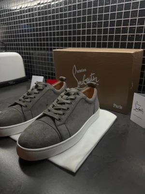 Gråa sneakers från Christian Louboutin - Snygga gråa sneakers från Christian Louboutin i mocka med vita sulor och klassisk låg profil. Skorna har gråa skosnören, diskreta sömmar och en stilren design som passar till det mesta. Insidan är fodrad i beige läder för extra komfort.