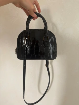 Svart croco handväska med axelrem - Snygg svart handväska i glansigt croco-mönstrat konstläder. Väskan har rundad form, två handtag och en lång justerbar axelrem. Guldiga metalldetaljer ger en extra touch. Perfekt för dig som vill ha en trendig och stilren accessoar.