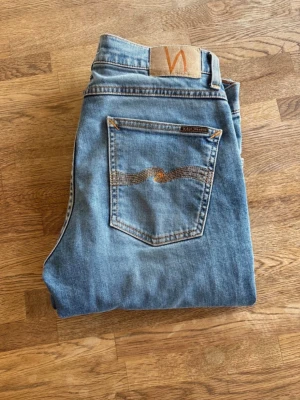 Ljusblå jeans från Nudie Jeans - Säljer ett par klassiska ljusblå jeans från Nudie Jeans med ikonisk vågsöm på bakfickan och läderpatch i midjan. Jeansen har rak passform och är tillverkade i slitstark denim. Perfekta för en avslappnad och trendig look.