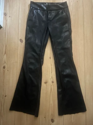 Svarta skinnbyxor med utsvängda ben - Snygga svarta skinnbyxor med utsvängda ben och klassisk midja. Byxorna har en slät finish och stängs med knapp och dragkedja. Perfekta för dig som vill ha en cool och edgy look. Köpta på berska med klippta ben för att de va för långa. Även klippt som ni ser på bilden men inget som syns eller påverkar än när man har på sig de.
