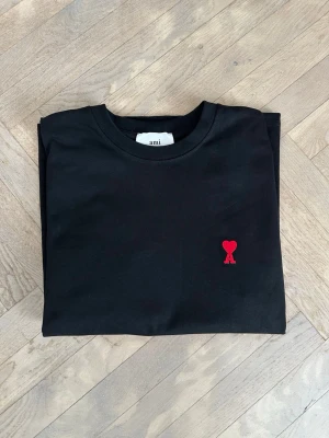 Svart t-shirt från Ami Paris - Säljer en svart t-shirt från Ami Paris med klassisk passform och korta ärmar. På bröstet finns ett broderat rött hjärta med bokstaven A, vilket är varumärkets ikoniska detalj. Perfekt för dig som gillar stilrena plagg med en twist. Priset är inte hugget i sten, så hör gärna av dig vid intresse!