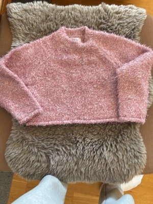 Rosa fluffig stickad tröja - En supermysig rosa stickad tröja med fluffig textur och långärmad design. Tröjan har en hög hals och är tillverkad i ett mjukt, glansigt material som ger en cool och trendig look. Perfekt för dig som gillar att sticka ut och vill ha något riktigt cozy.