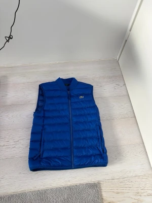 Blå dunväst från Lacoste - Snygg blå dunväst från Lacoste med klassisk quiltad design och dragkedja framtill. Västen har två sidofickor och den ikoniska Lacoste-loggan på bröstet. Perfekt att slänga över en t-shirt för en cool look. Size m but fit s