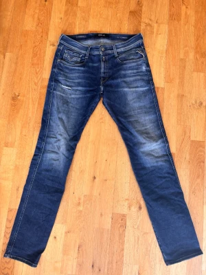 Blå slim fit jeans från Replay - Snygga blå slim fit jeans från Replay med klassisk femficksdesign och diskreta slitningar på framsidan. Jeansen är i storlek w31 l34. Bild 3 är en lagning av ett hål gjord till en snygg slitning.