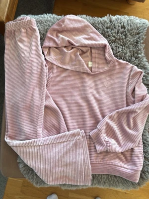 Rosa ribbad loungewear-set med huva - Mysigt loungewear-set i ljusrosa ribbad velour med huva och långa ärmar. Setet består av en hoodie och matchande byxor med resår i midjan och vid benslut. Liten broderad hjärt-detalj på bröstet. Perfekt för chill dagar hemma.