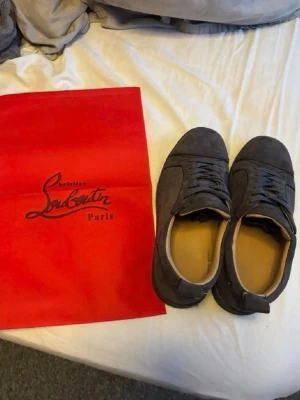 Gråa sneakers från Christian Louboutin - Snygga gråa sneakers från Christian Louboutin i mocka med klassisk låg siluett och matchande grå snörning. Skorna har en stilren design och ikonisk röd detaljpåse medföljer. Perfekta för dig som vill ha något exklusivt och trendigt.