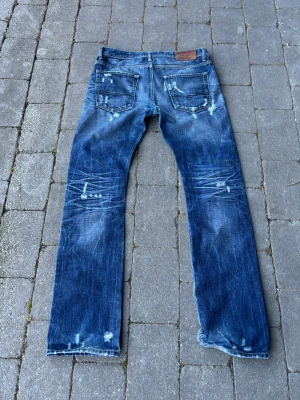 Blå slitna jeans med raka ben - Säljer ett par blå jeans med raka ben och tydliga slitningar samt distressed detaljer framtill och baktill. Jeansen har klassisk femficksdesign och är tillverkade i denim. Perfekta för en avslappnad och trendig look.