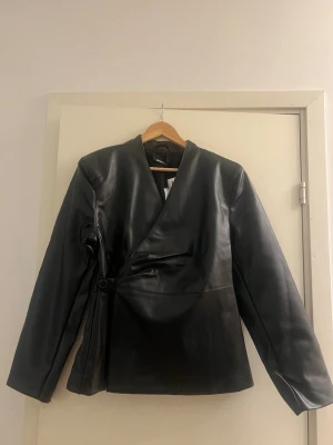Svart PU-kavaj från Gina Tricot - Snygg svart kavaj i PU-material från Gina Tricot. Modellen har omlottknäppning med bälte i sidan och en stilren, minimalistisk look. Perfekt för dig som vill ha en edgy och trendig vibe till din outfit.